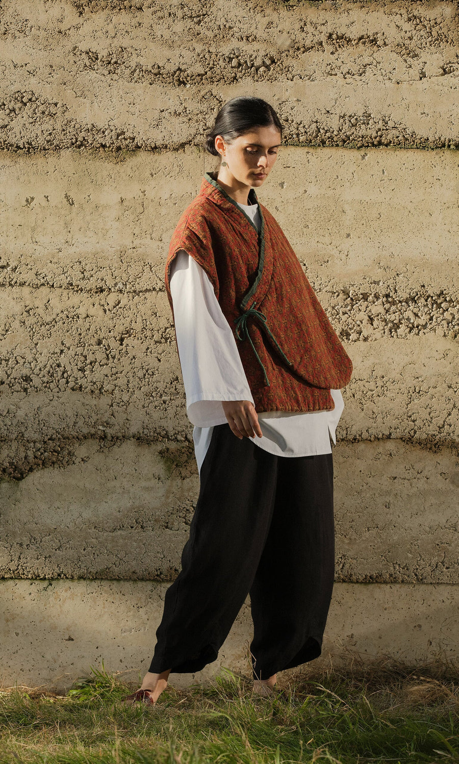 Plümo- A person stands outdoors in loose black pants, a white shirt, and the Ansel Wrap Jacket—a lightly quilted red vest.
