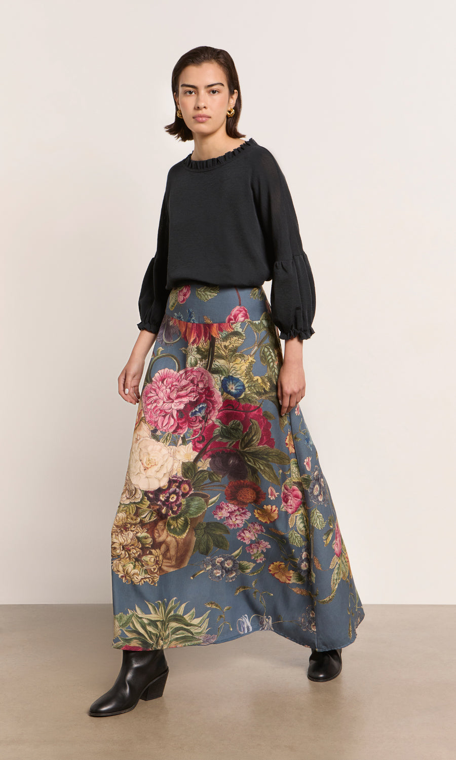 Plümo  Woman in black top and Aubrey Skirt, wearing black boots, stands against a plain light wall.