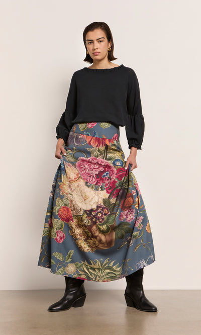 Plümo  Woman in a black blouse and the Aubrey Skirt stands against a plain background, wearing sustainable black boots.