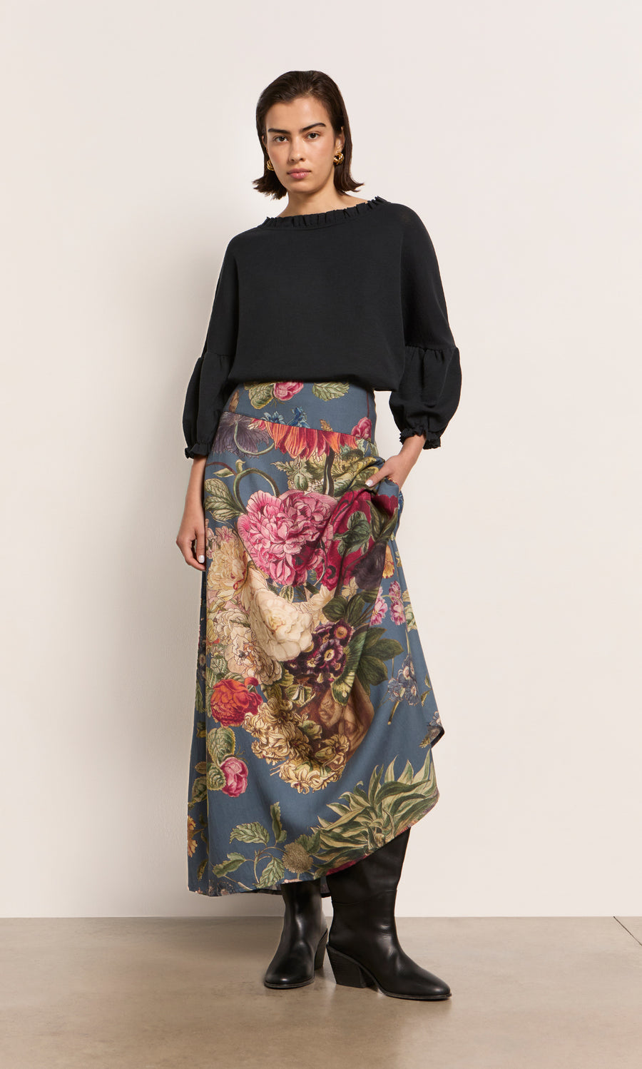 Plümo  Woman in a black top, Aubrey Skirt with organic floral print, and black boots stands by a pale wall.
