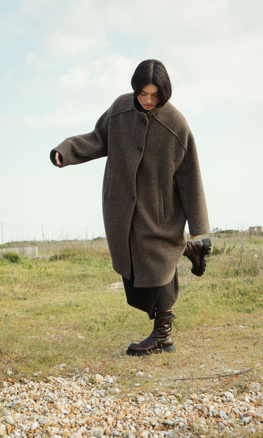 Plümo- A woman in the Babette Coat balances on one foot outdoors on a grassy, rocky field under a cloudy sky.
