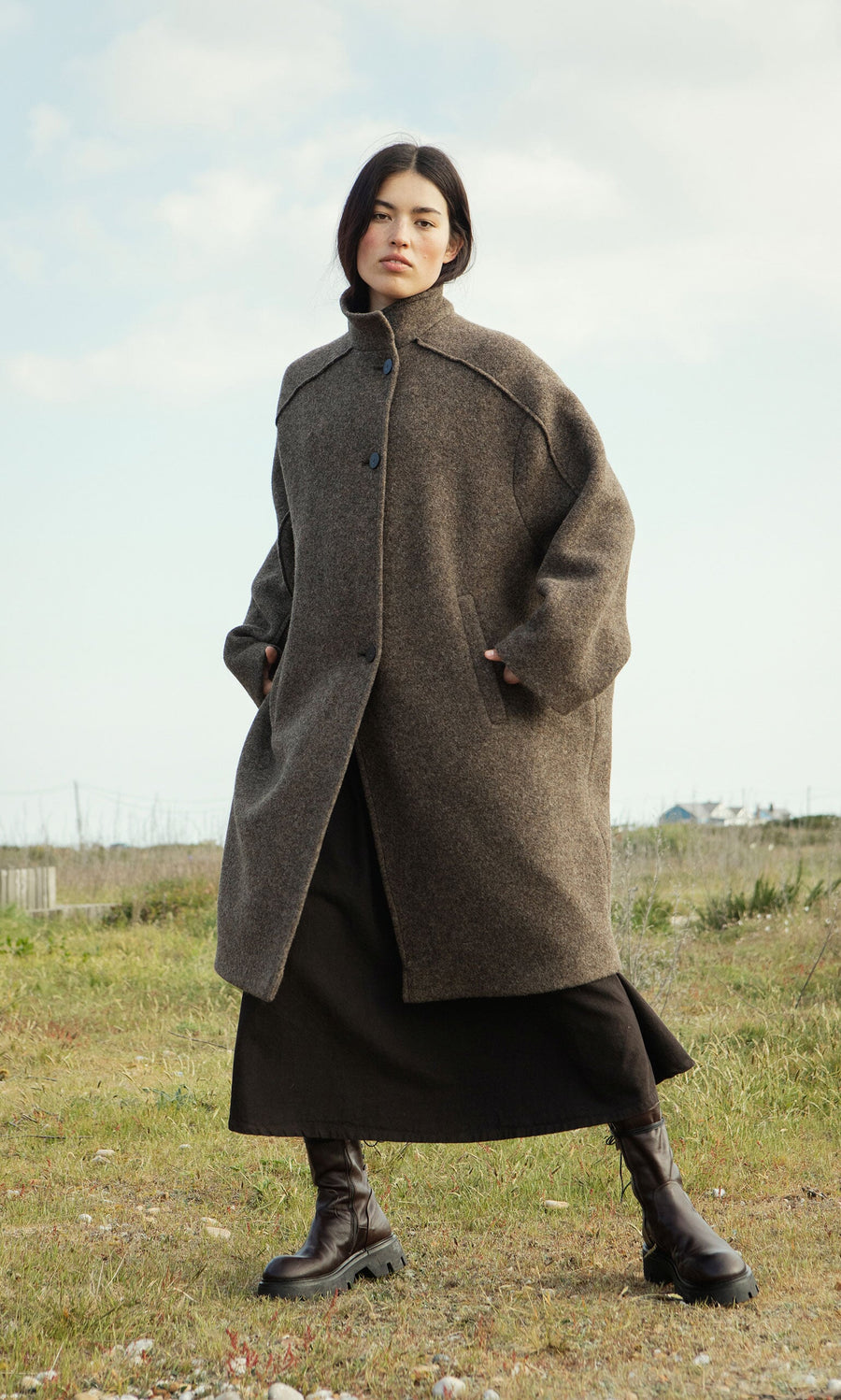 Plümo- A person in the Babette Coat, black boots, and a dark skirt stands on grass beneath a cloudy sky.