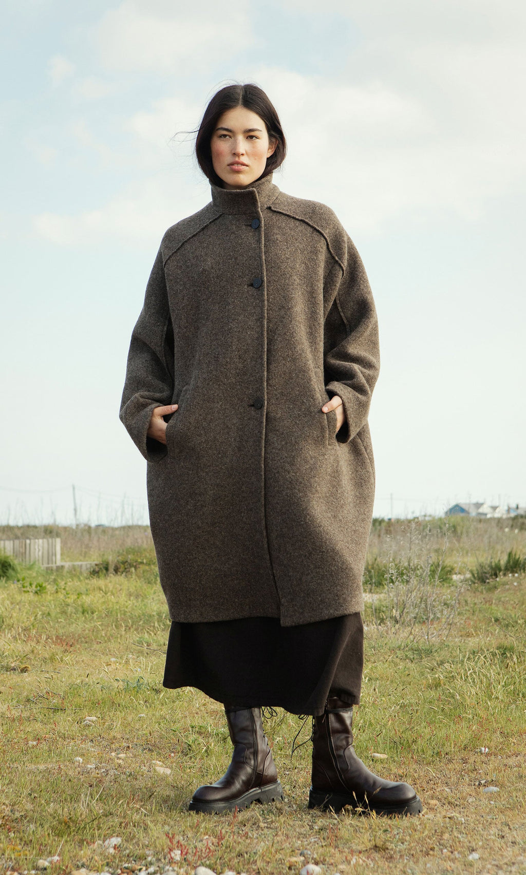 Plümo- Woman wears the Babette Coat and boots, standing on grass under a partly cloudy sky.