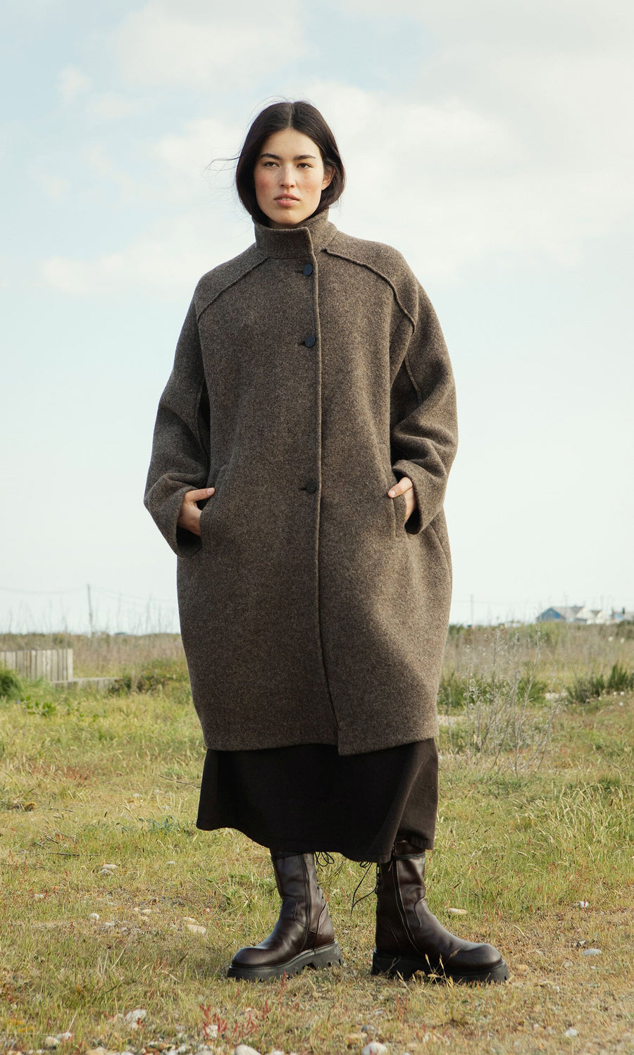 Plümo- Woman wears the Babette Coat and boots, standing on grass under a partly cloudy sky.