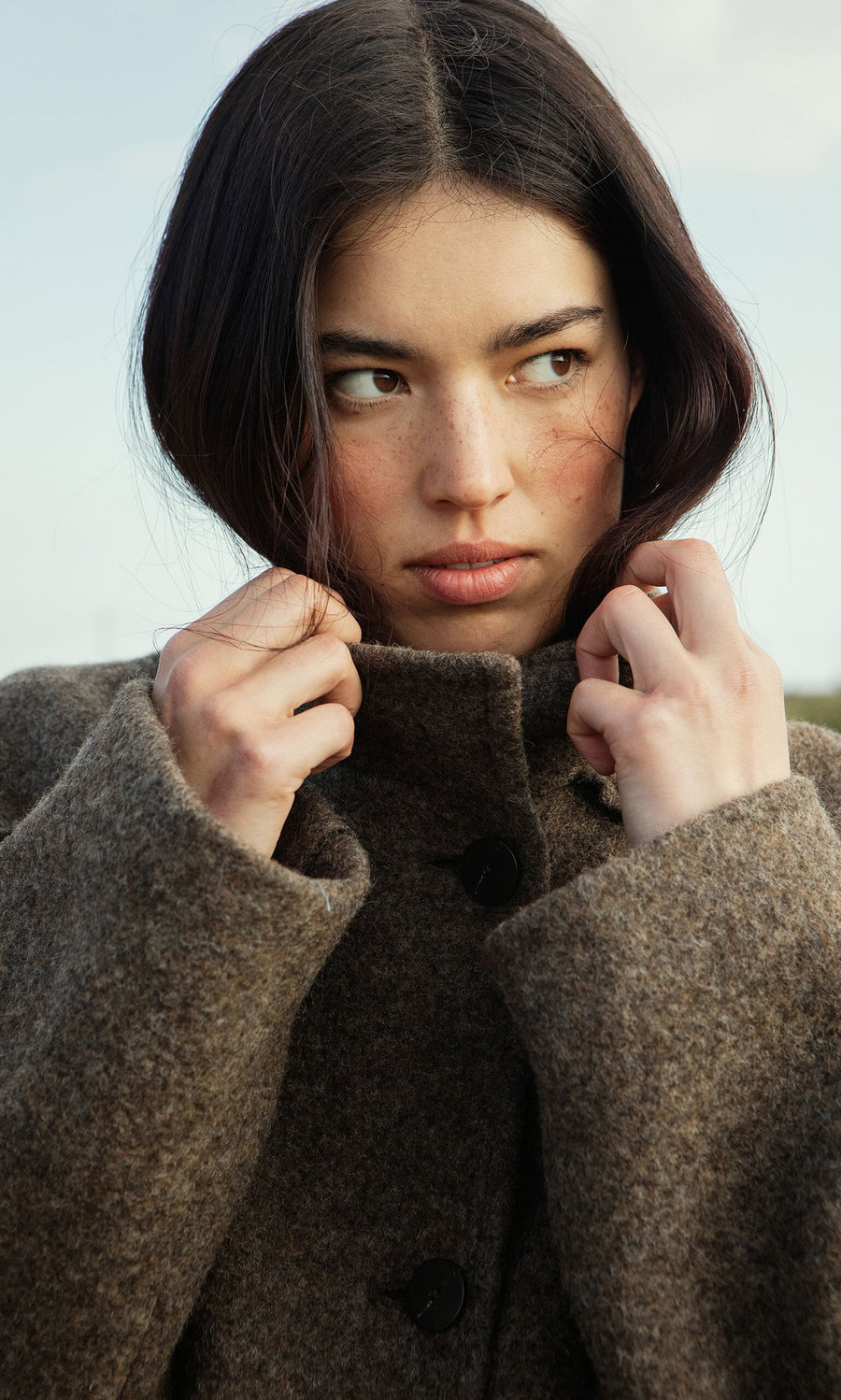 Plümo- Woman with dark hair wears the Babette Coat in felted Italian wool, collar up, looking to the side seriously.