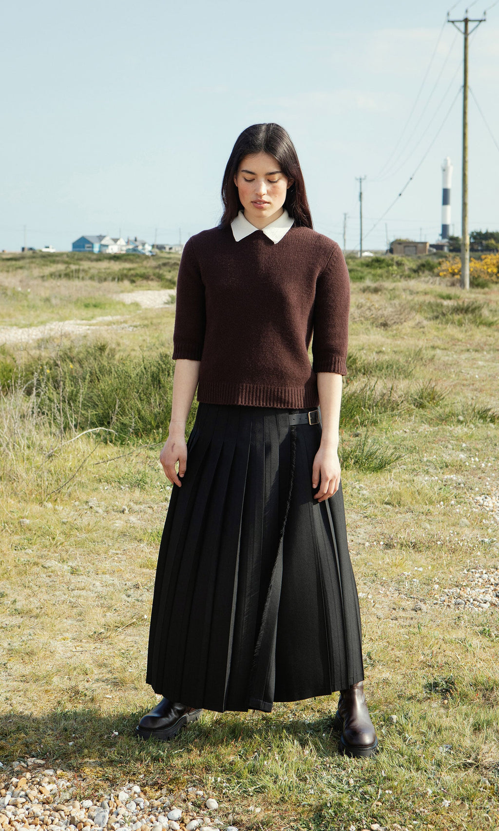 Plümo- A woman stands on grass wearing the boxy fit Bancroft Jumper, a white shirt, pleated skirt, and brown boots.