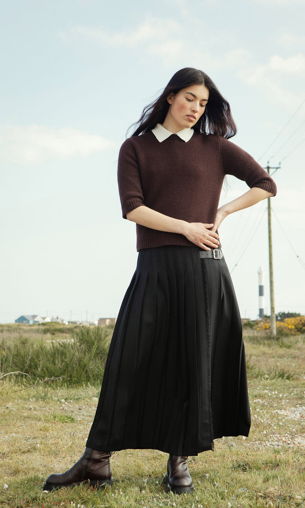Plümo- Woman in a Bancroft Jumper and black pleated skirt stands outdoors with a lighthouse and buildings in the background.