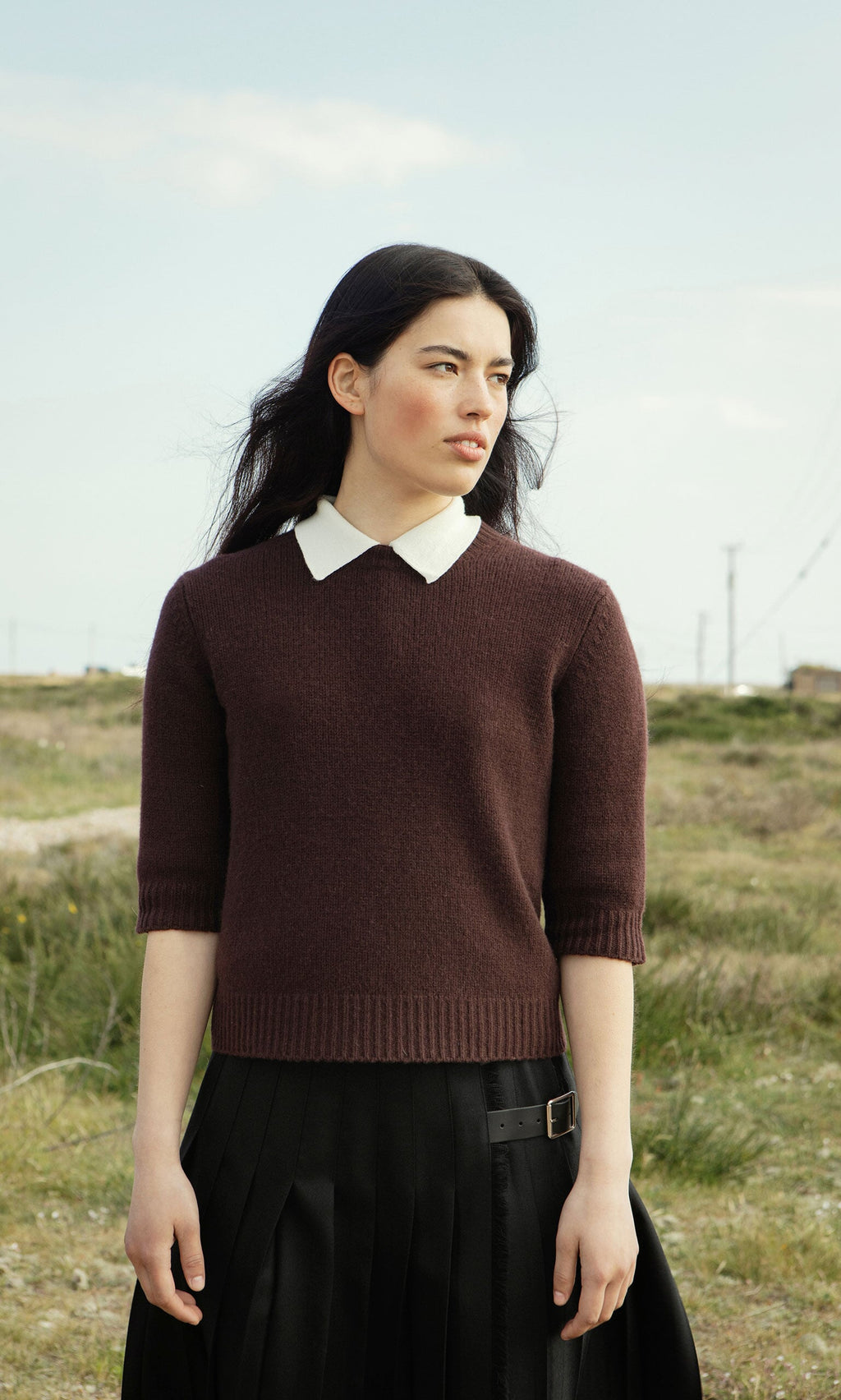 Plümo- A woman in a supersoft Bancroft Jumper and black skirt stands outside on a grassy field, looking to the side.