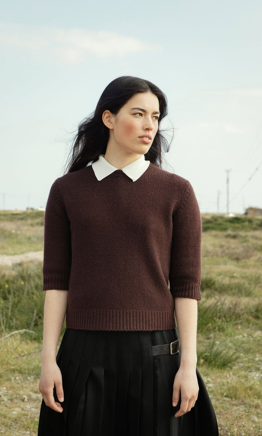 Plümo- A woman in a supersoft Bancroft Jumper and black skirt stands outside on a grassy field, looking to the side.