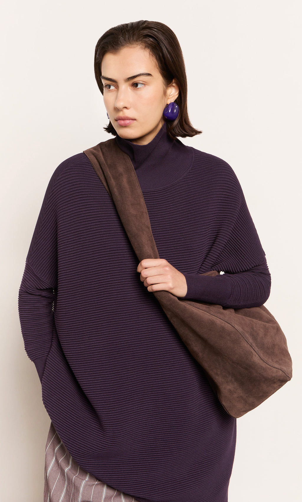 Plümo  Woman in a purple turtleneck, large earrings, and chocolate brown suede bag poses against a plain background.