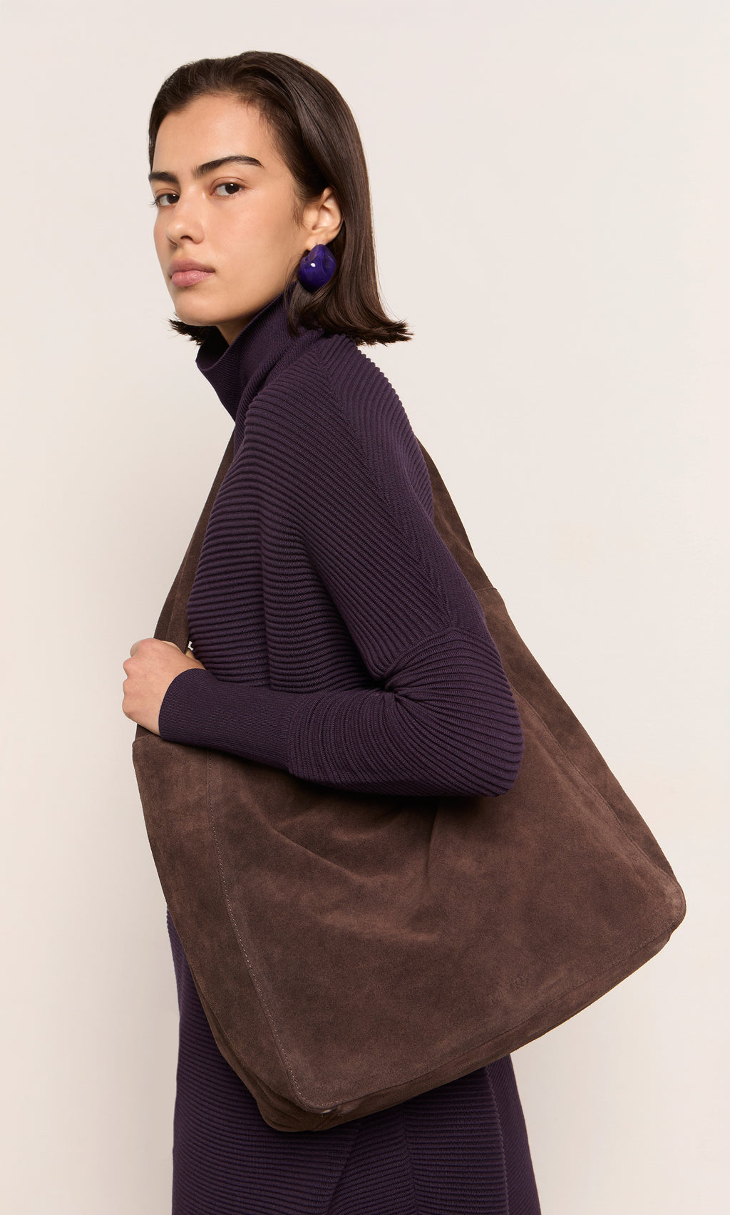 Plümo  Woman in a purple ribbed dress holds a Bento Suede Bag in chocolate brown, looking confidently at the camera.