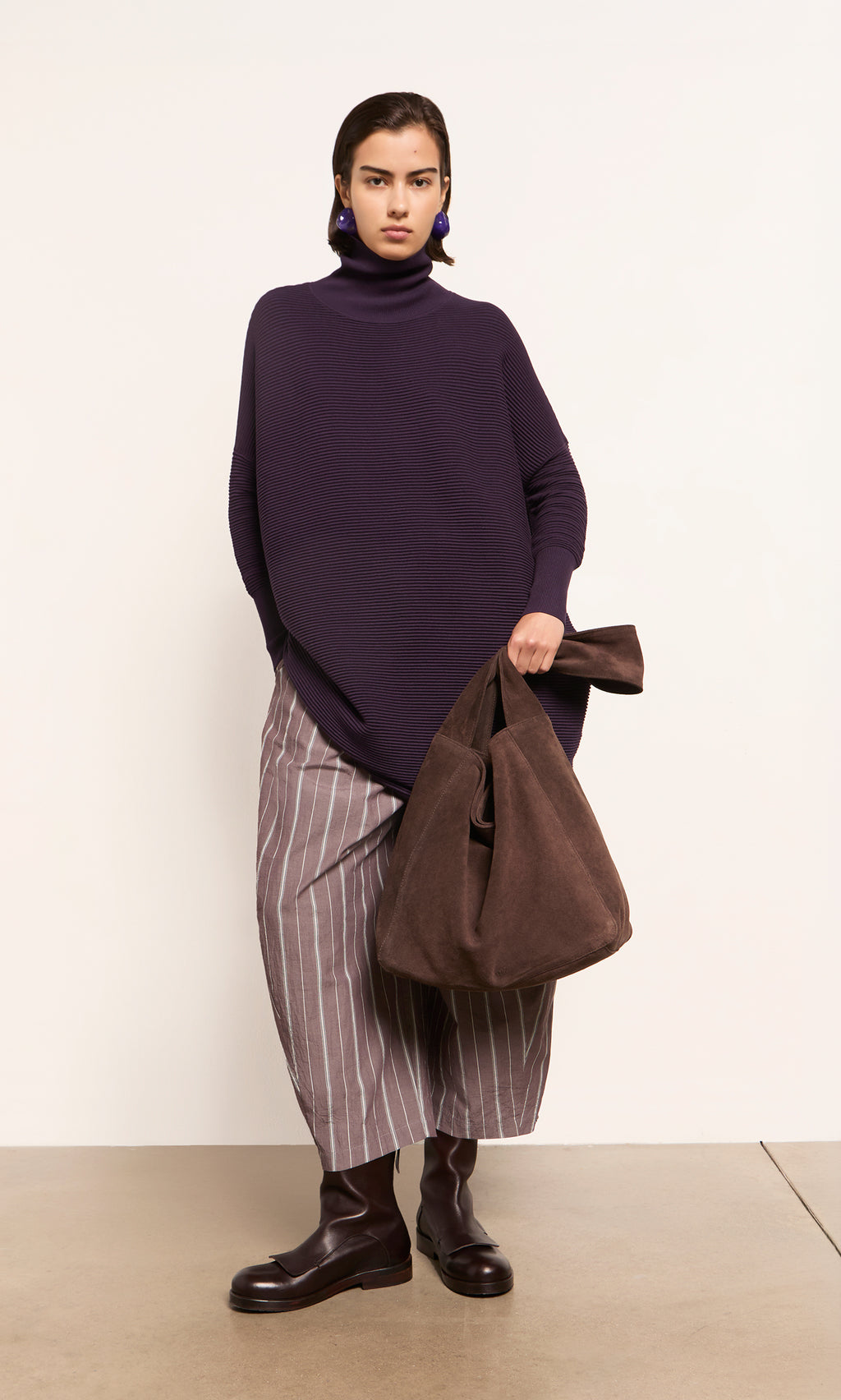 Plümo  Woman with a Bento Suede Bag, in a purple turtleneck, striped skirt, and brown boots stands by a white wall.