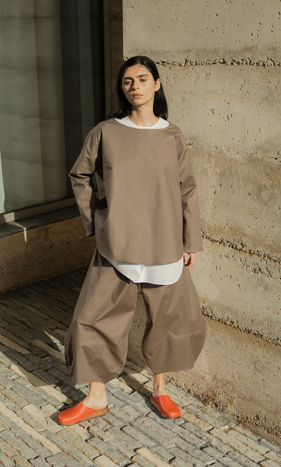 Plümo- A woman in loose brown pants, orange shoes, and a Beranda Stripe Top stands against a concrete wall.