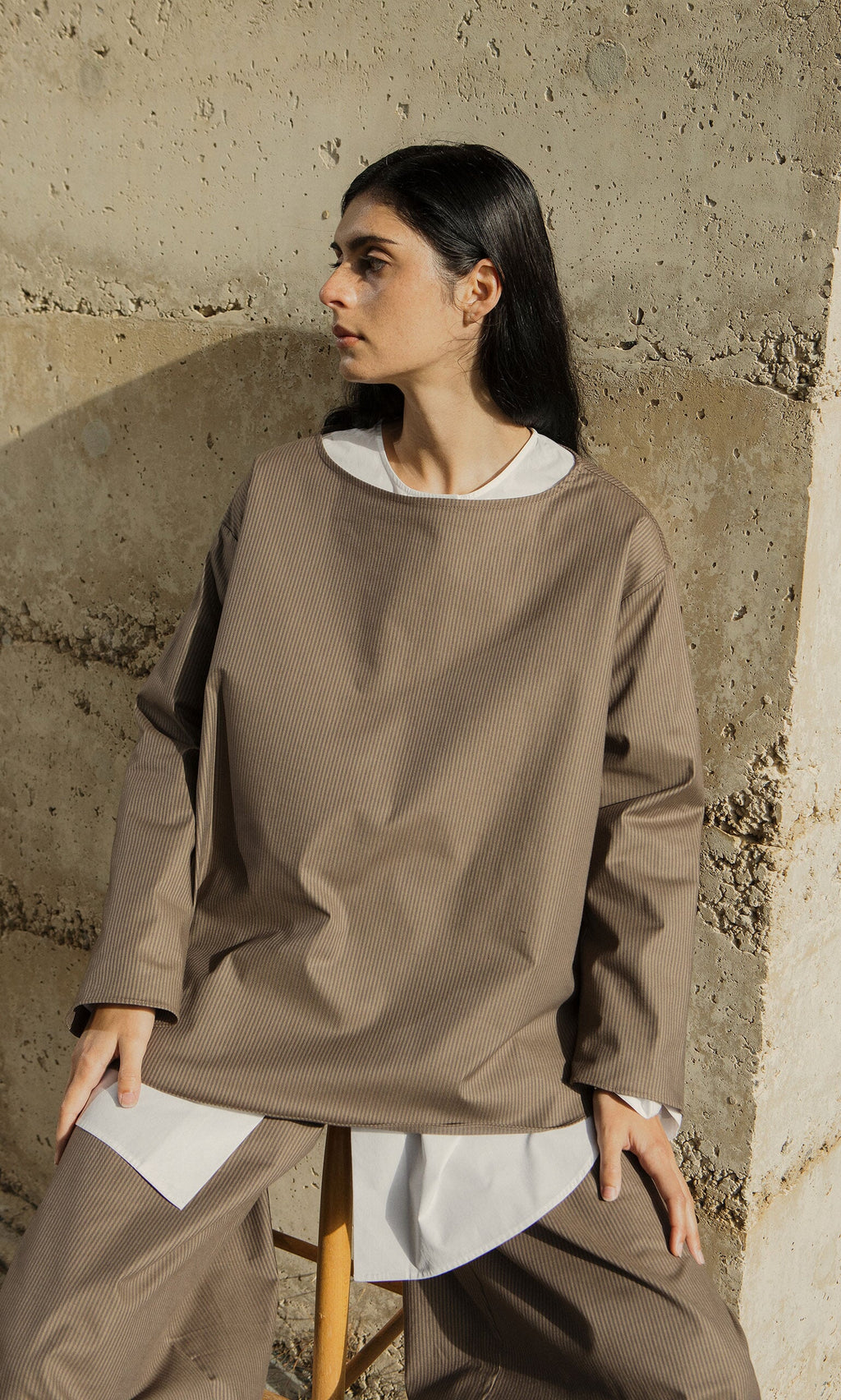 Plümo- A person in the Beranda Stripe Top sits on a stool by a textured concrete wall, looking to the side.
