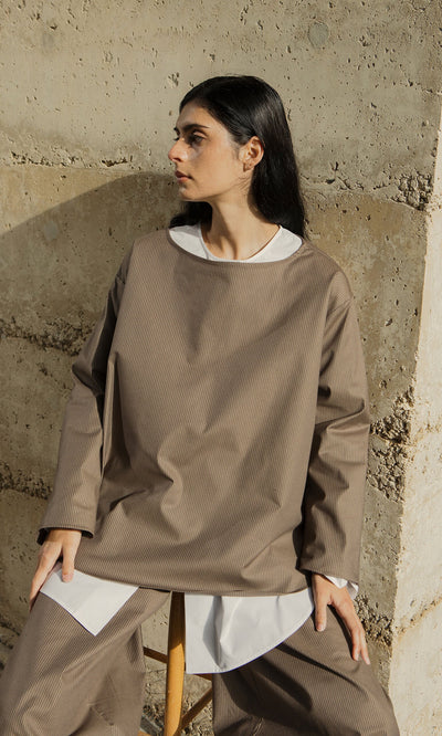 Plümo- A person in the Beranda Stripe Top sits on a stool by a textured concrete wall, looking to the side.