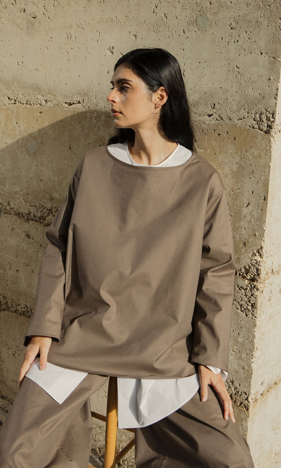 Plümo- A person in the Beranda Stripe Top sits on a stool by a textured concrete wall, looking to the side.