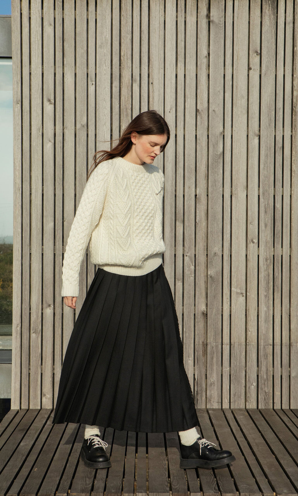 Plümo- Woman in a Berg Aran Sweater and black pleated skirt stands on a wooden deck with slatted wood wall behind her.