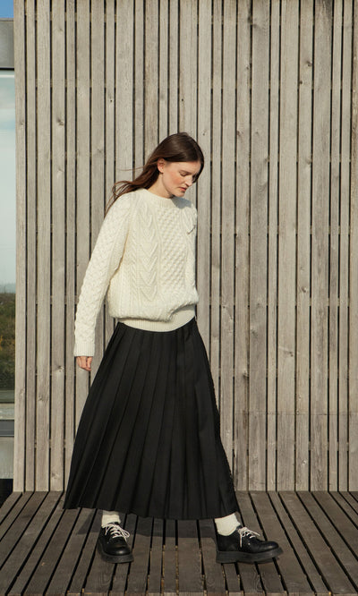 Plümo- Woman in a Berg Aran Sweater and black pleated skirt stands on a wooden deck with slatted wood wall behind her.