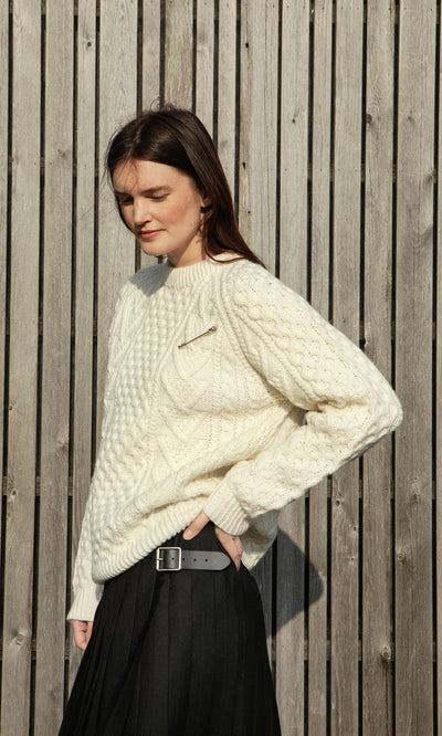 Plümo- Woman in a Berg Aran Sweater and black skirt stands by a wooden fence, gazing to the side.