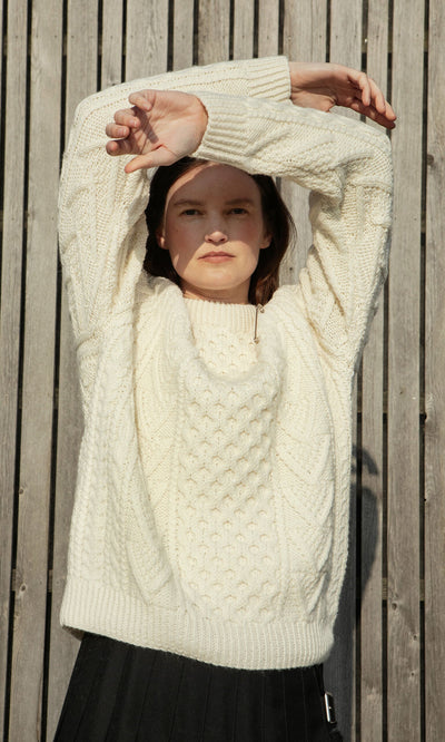 Plümo- Person in a Berg Aran Sweater stands before a wooden fence, arms raised, sunlight shining on their face.