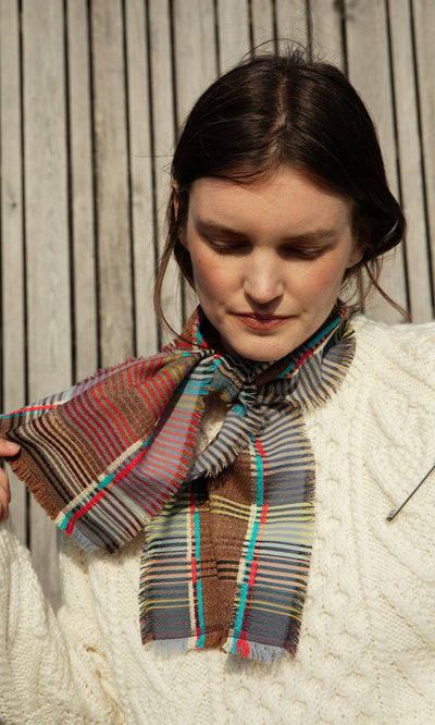Plümo- Woman in a Berg Aran Sweater with a colorful plaid scarf, looking down beside a wooden wall.