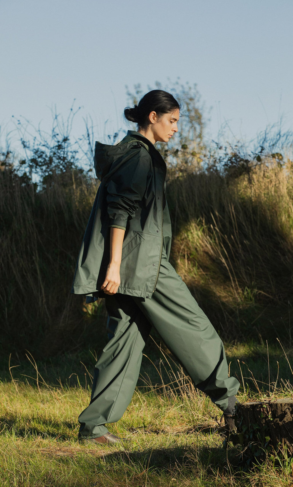 Plümo- Wearing a Berghart Raincoat, someone walks through grassy terrain on a sunny day amid tall grass and trees.