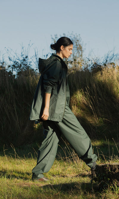 Plümo- Wearing a Berghart Raincoat, someone walks through grassy terrain on a sunny day amid tall grass and trees.