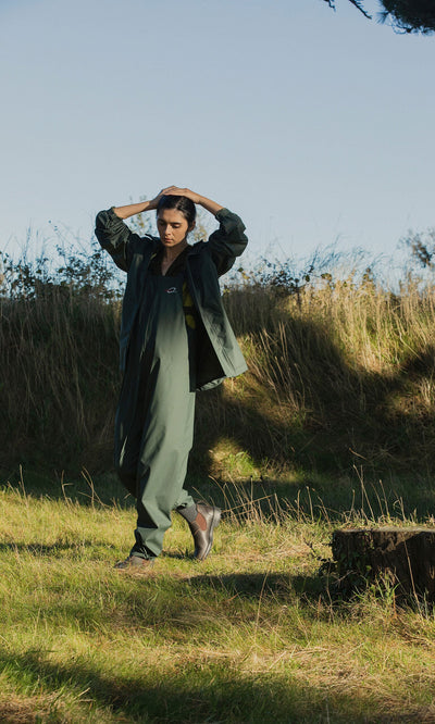 Plümo- A person in a green Berghart Raincoat and pants stands on grass outdoors in sunlight near bushes and trees.