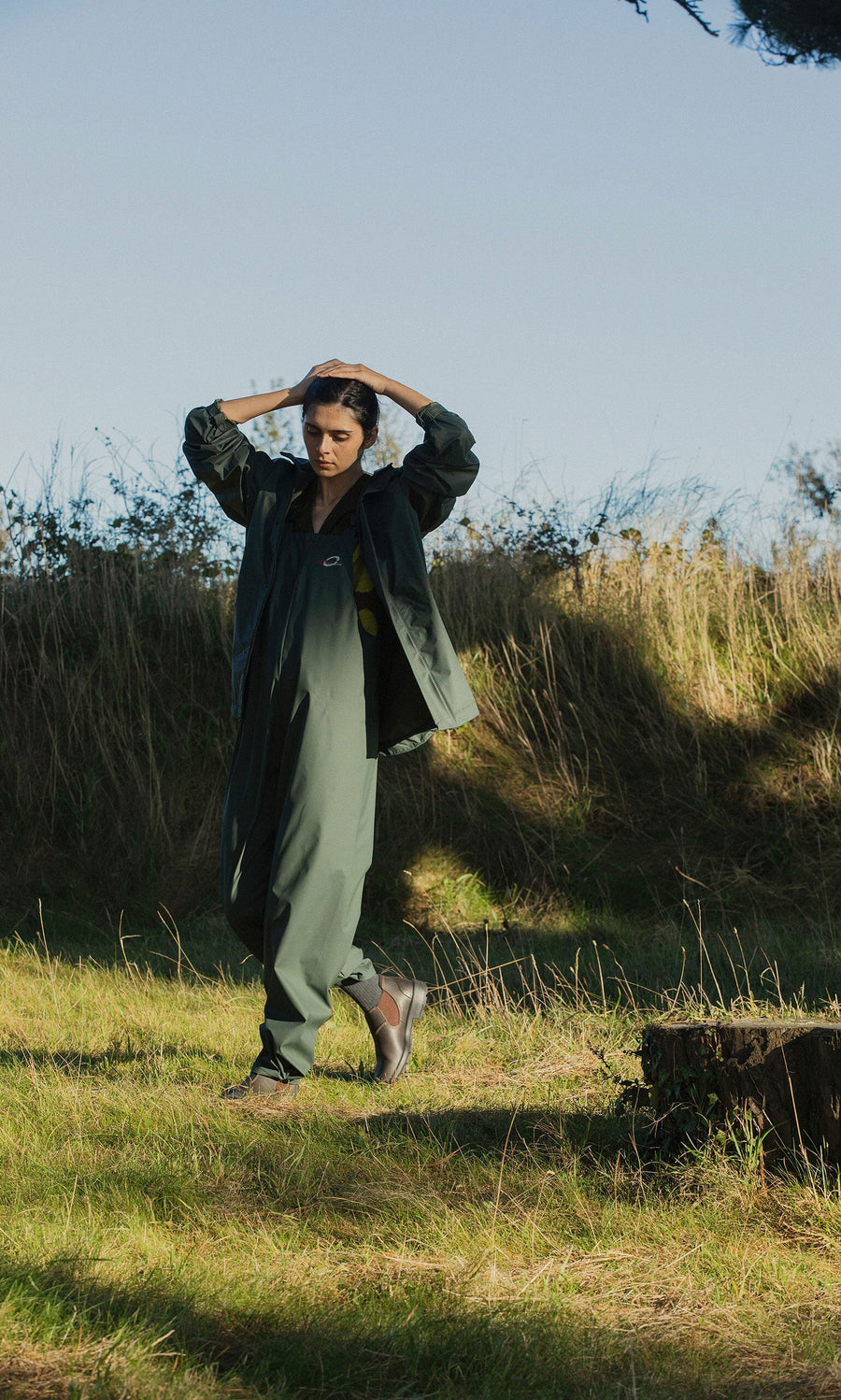 Plümo- A person in a green Berghart Raincoat and pants stands on grass outdoors in sunlight near bushes and trees.