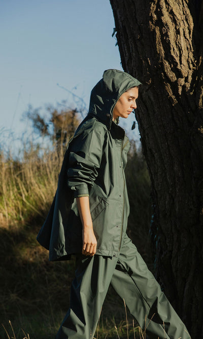 Plümo- Person in a green Berghart Raincoat with hood walking outdoors by a large tree in natural sunlight.