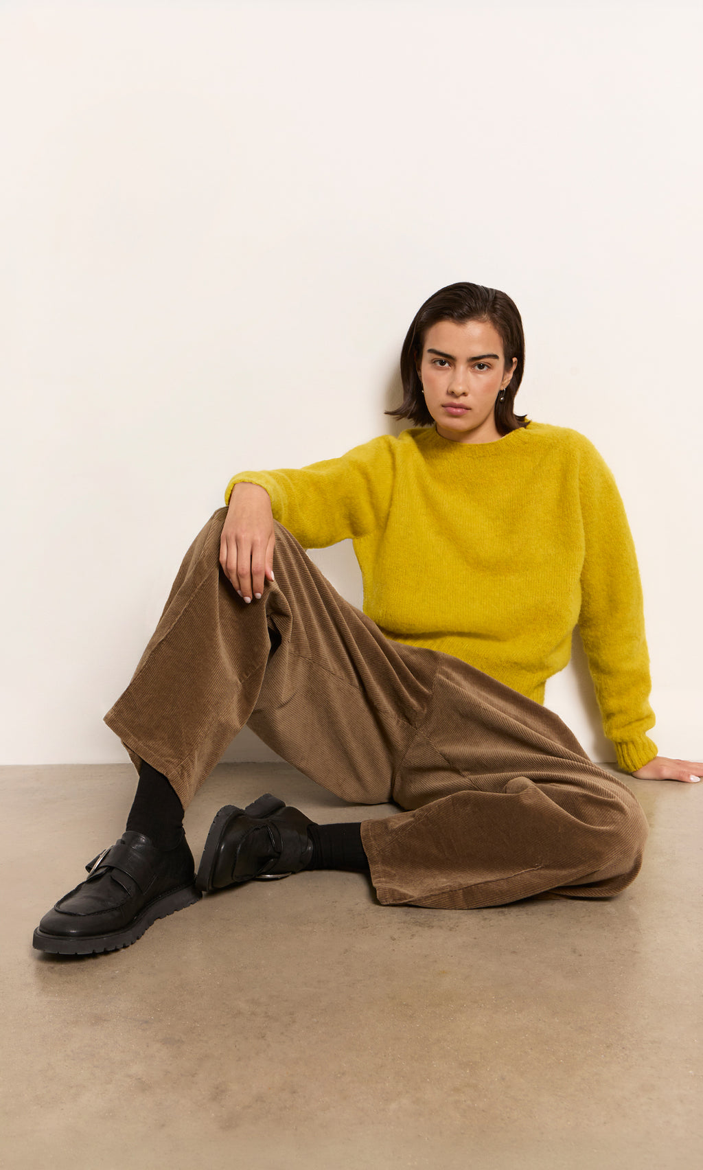 Plümo  Person in a yellow sweater and brown pants wears Black Creepers, sitting on the floor and looking at the camera.