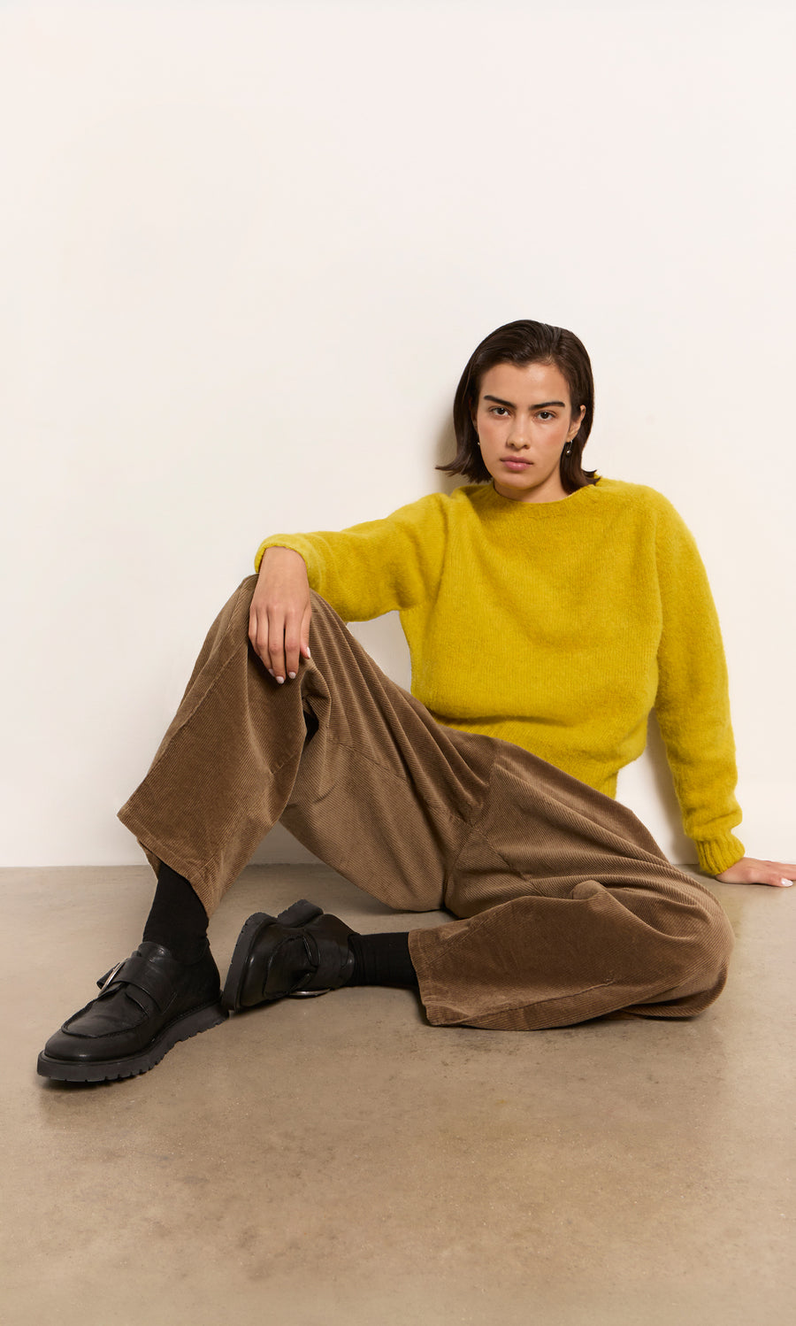 Plümo  Person in a yellow sweater and brown pants wears Black Creepers, sitting on the floor and looking at the camera.