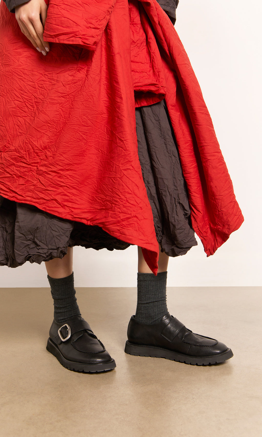 Plümo  Person in a red coat and Black Creepers with lugged rubber outsoles stands on a tan floor.
