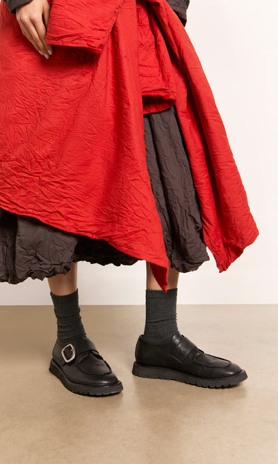 Plümo  Person in a red coat and Black Creepers with lugged rubber outsoles stands on a tan floor.