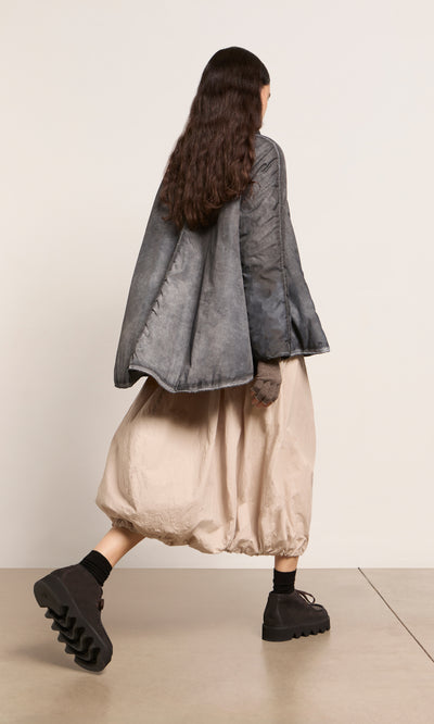 Plümo  Person with long dark hair in a Black Pepper Jacket, beige skirt, and black shoes, walking to the left.