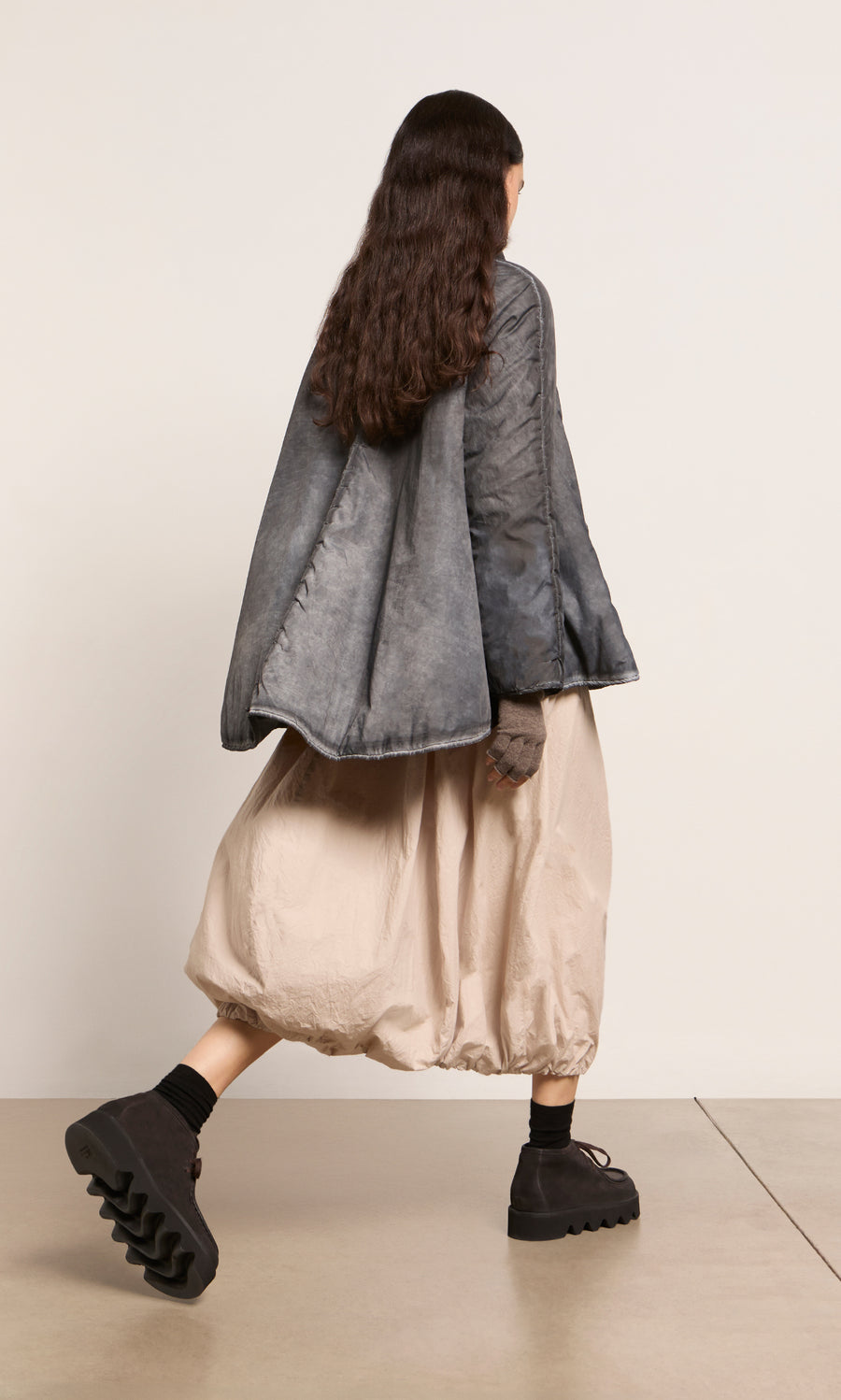 Plümo  Person with long dark hair in a Black Pepper Jacket, beige skirt, and black shoes, walking to the left.