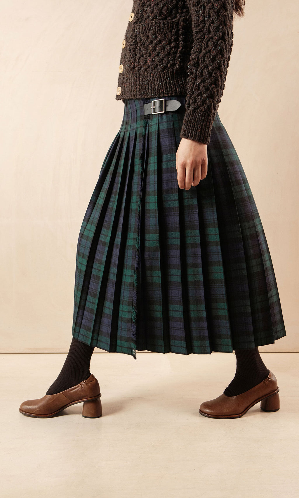 Plümo- Person wearing a Blackwatch Kilt, brown sweater, black socks, and brown heeled shoes while walking indoors.