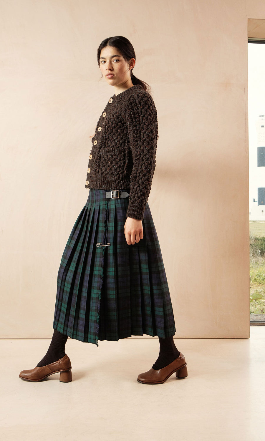 Plümo- Woman in a brown knit sweater and Blackwatch Kilt, walking indoors wearing brown shoes and socks.