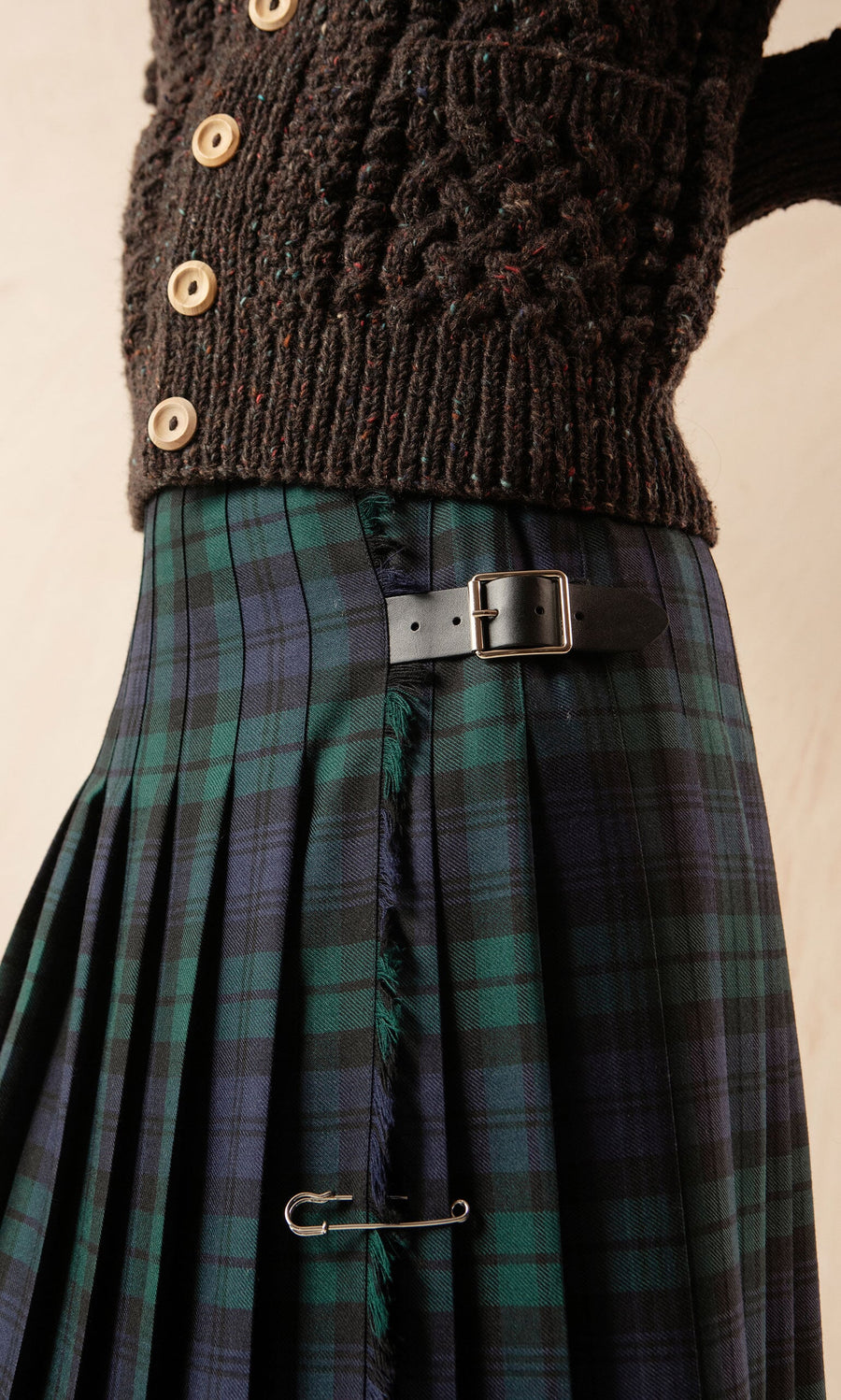 Plümo- Close-up of the Blackwatch Kilt, made in Ireland, styled with a belt, safety pin, and buttoned dark textured sweater.