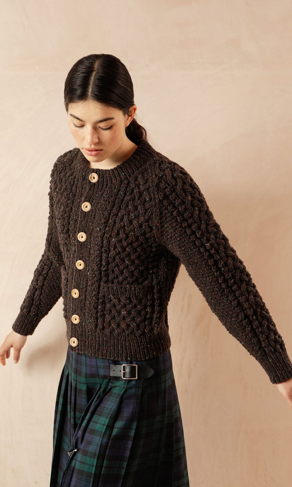 Plümo- Woman wears a dark knit cardigan and the Blackwatch Kilt, standing against a pale background.