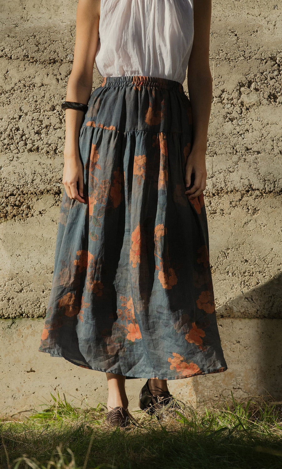 Plümo- A person in a sleeveless white top and the Bloom Skirt with a grey floral print and elasticated waist stands on grass.