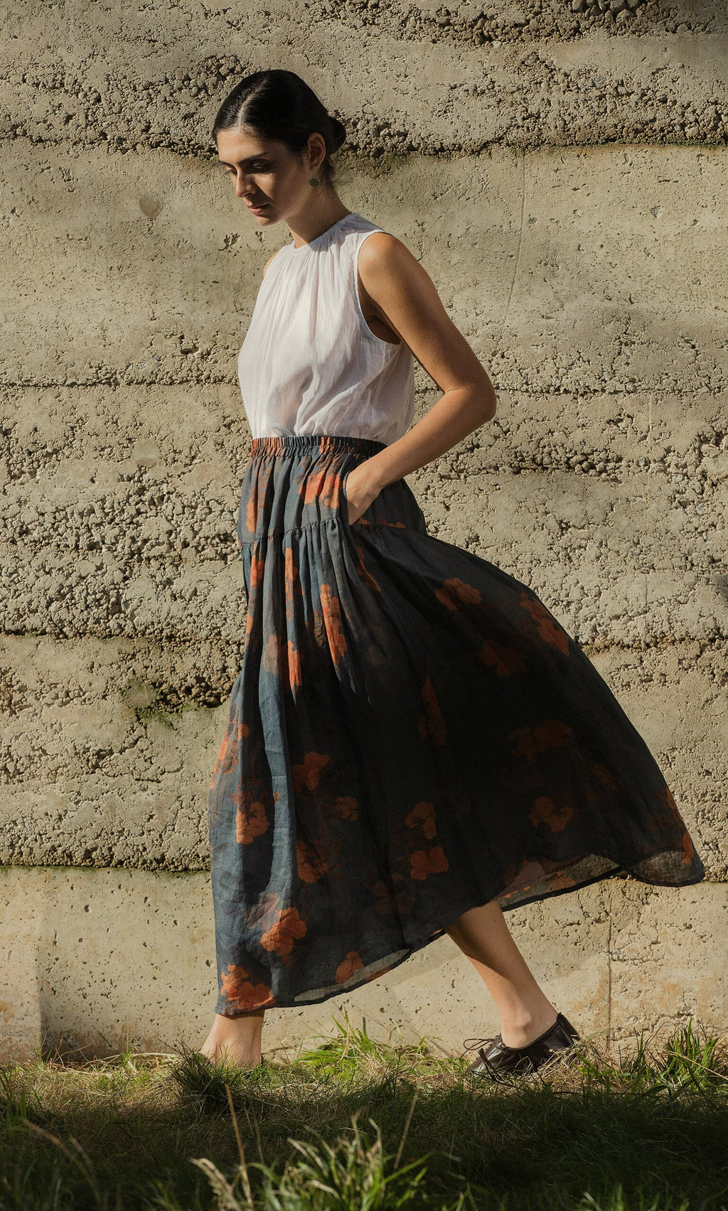 Plümo- Woman in a sleeveless white top and the Bloom Skirt walks by a textured concrete wall.