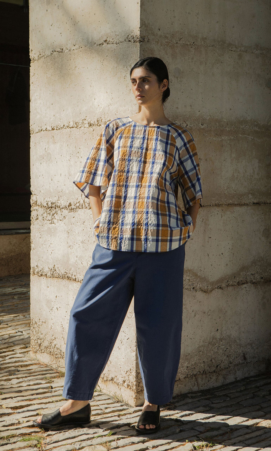 Plümo- A person in a Blue Madras Shirt and blue pants stands against a concrete wall as sunlight casts shadows.