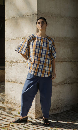 Plümo- A person in a Blue Madras Shirt and blue pants stands against a concrete wall, looking at the camera.
