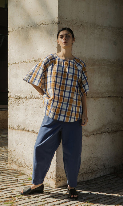 Plümo- A person in a Blue Madras Shirt and blue pants stands against a concrete wall, looking at the camera.