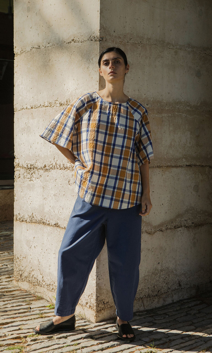 Plümo- A person in a Blue Madras Shirt and blue pants stands against a concrete wall, looking at the camera.