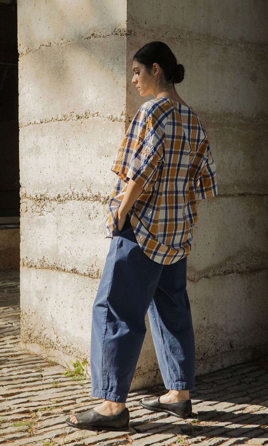 Plümo- Person outdoors in a Blue Madras Shirt, boxy fit, loose blue pants, and sandals, facing away.