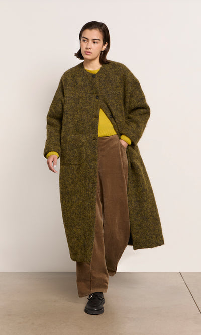Plümo  Person in a Borage Coat, yellow sweater, brown pants, and black shoes stands against a plain background.