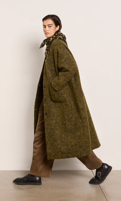 Plümo  Person in a long-line mossy green Borage Coat, brown pants, black shoes, and scarf walks by a plain light wall.