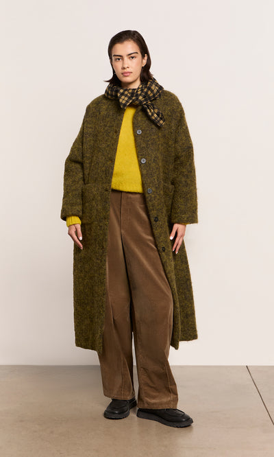 Plümo  Person wearing a long green coat, yellow sweater, brown pants, Quadrant Necktie, and black shoes indoors.